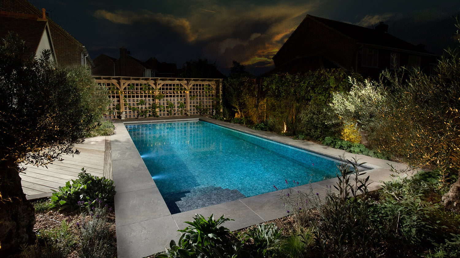 A swimming pool at night. Blue water with grey coping stones around. A fence at the back and lots of garden foliage around.