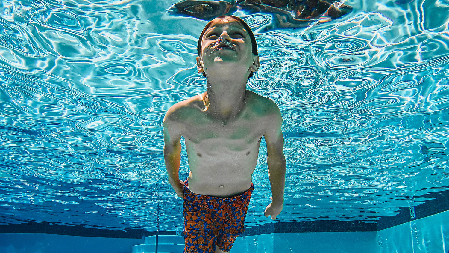 child swimming underwater in swimming pool. Child has eyes open and blowing air bubbles from nose. 