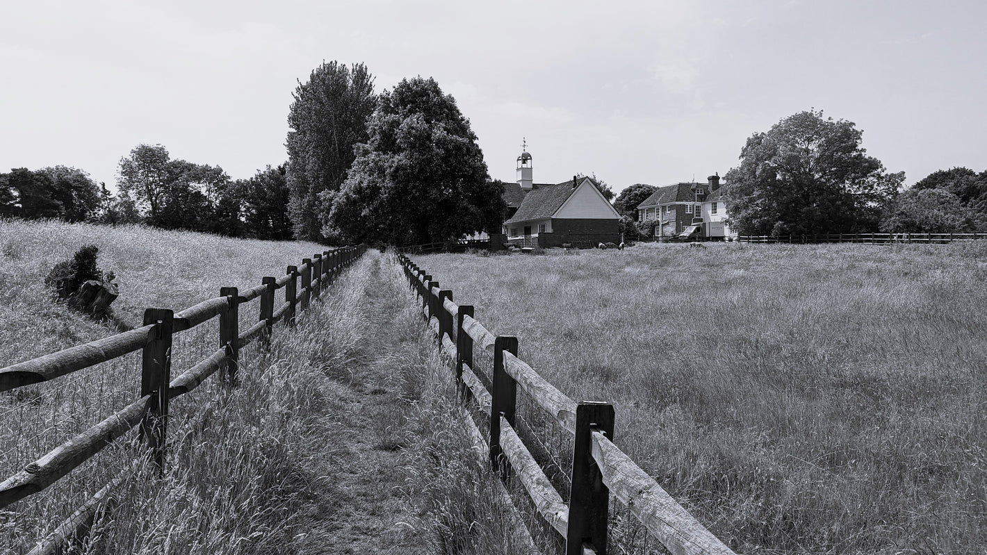 english countryside in black and white. Beautiful field with wooden fence lining path. Farmhouse in the background with trees to the left and right.