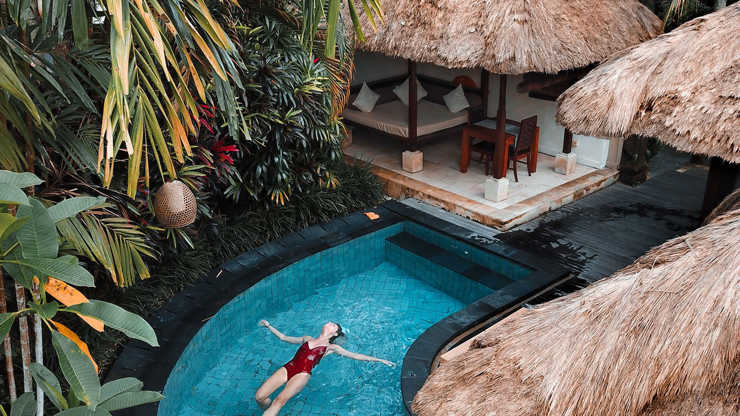 tropical pool with grass huts next to jungle foilage. Woman in red costume floating on her back relaxing in the pool.