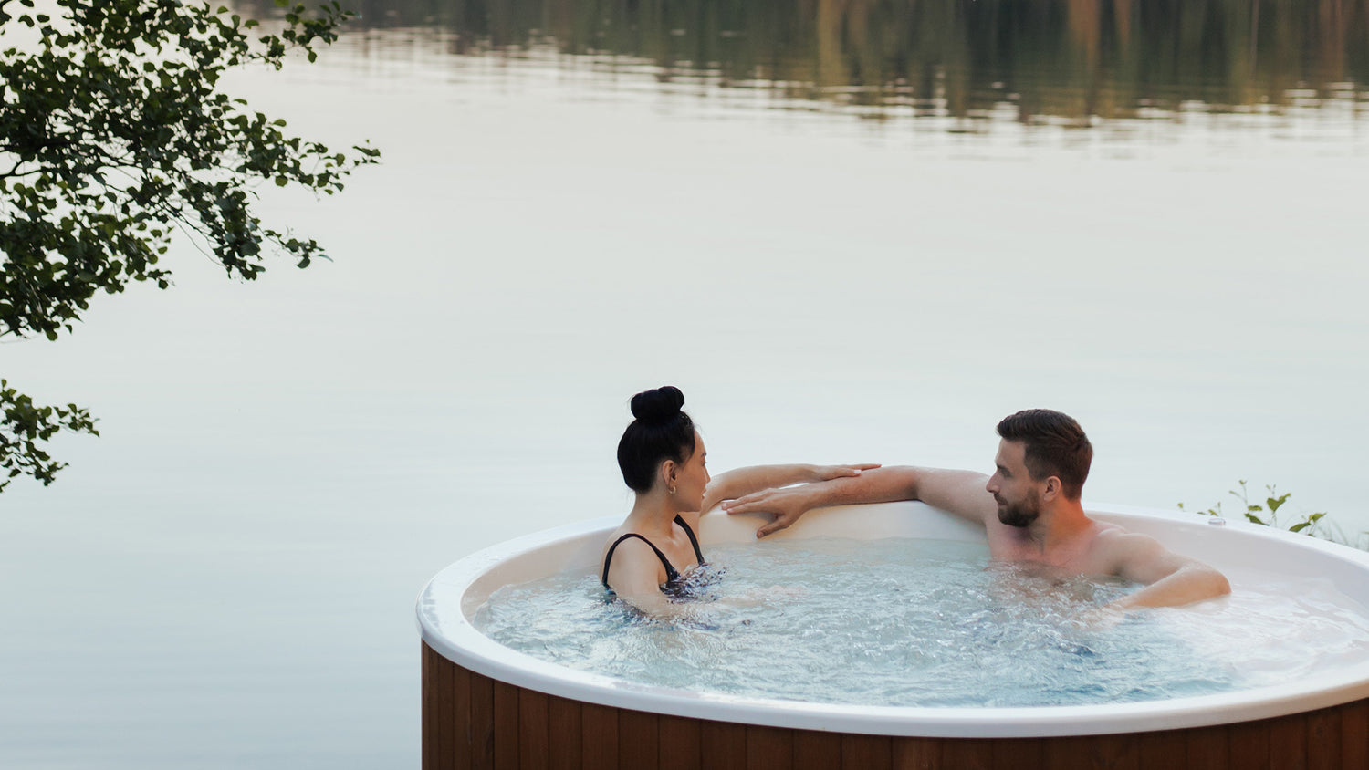 Round Hot Tub in the foreground, in front of a lake. Small tree to left of image. Couple relaxing and talking in hot tub.