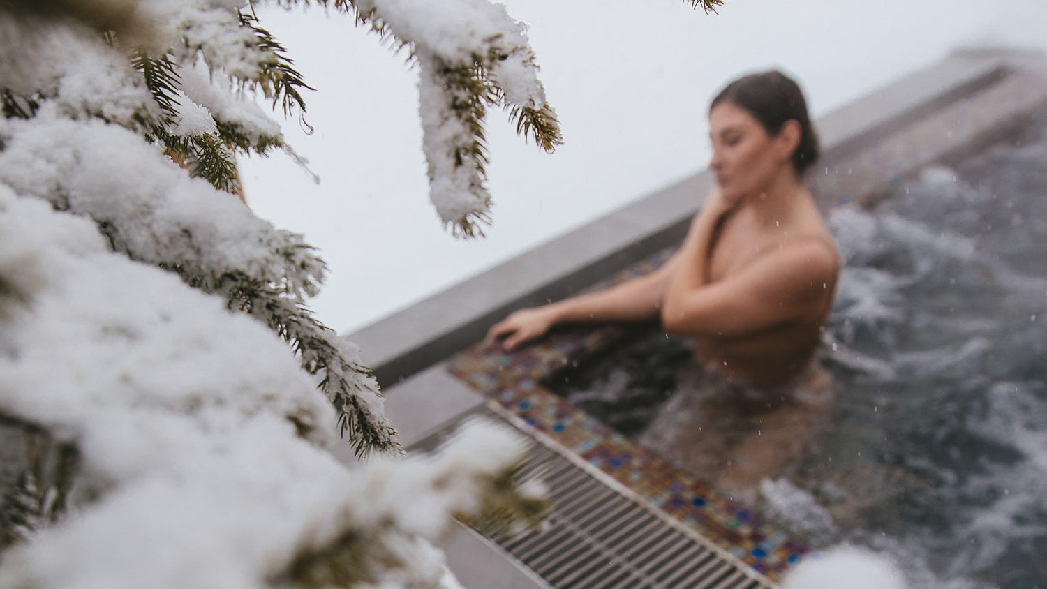 woman in hot tub in the winter. She is holding her shoulder. Snowy pine tree in foreground on left hand side.