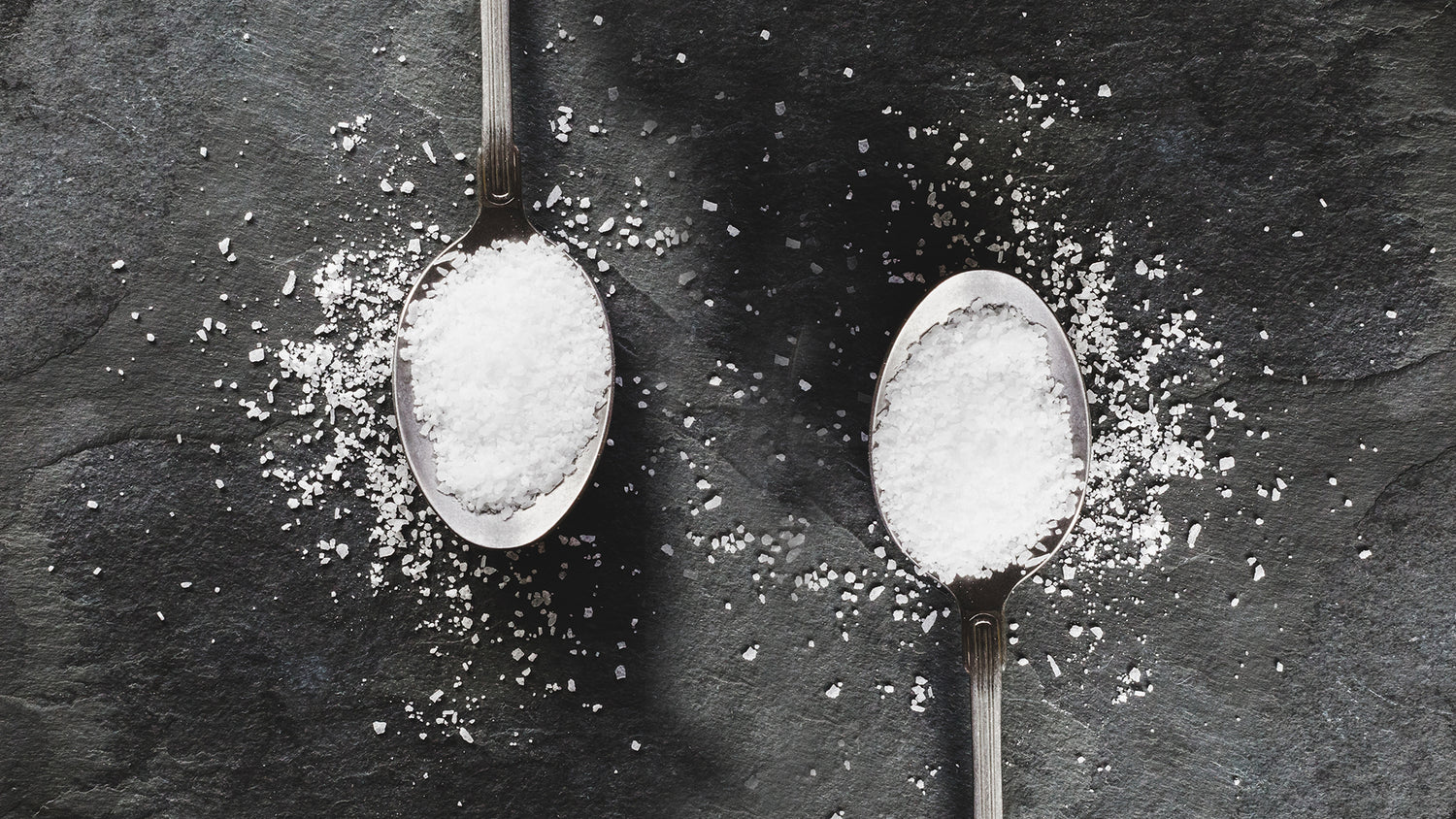 two teaspoons with white salt crystals in them on slate background. Sprinkling of salt around the spoons.
